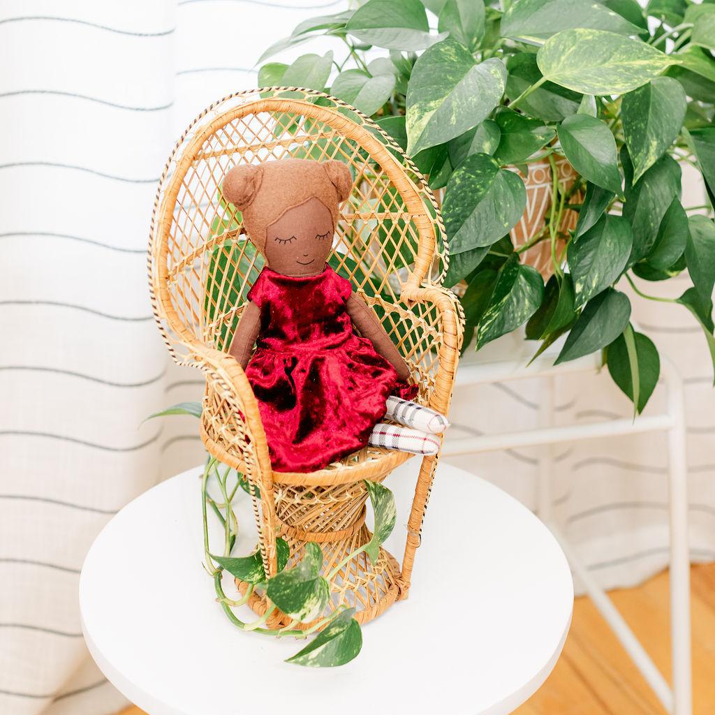 A doll with a maroon velvet dress presented sitting in a wicker chair against a backdrop of green plants.