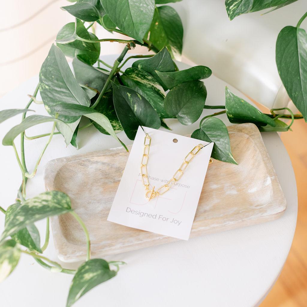 A gold-colored paperclip toggle necklace laid out on a white surface with green leaves in the background.