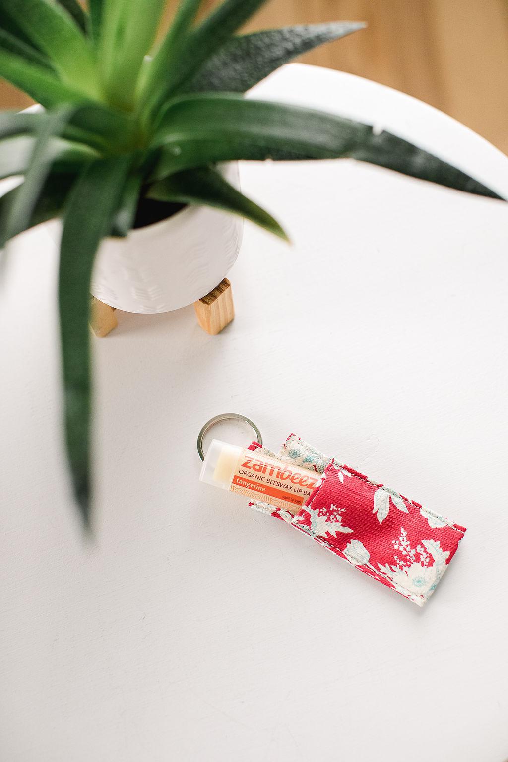 Lip balm with orange design in a red floral fabric sleeve on a keychain sitting on a white table with a plant.