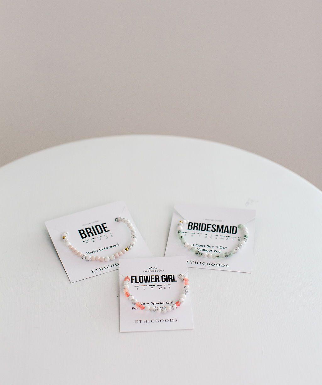 Three beaded bracelets on white cards on a white table. One card says &