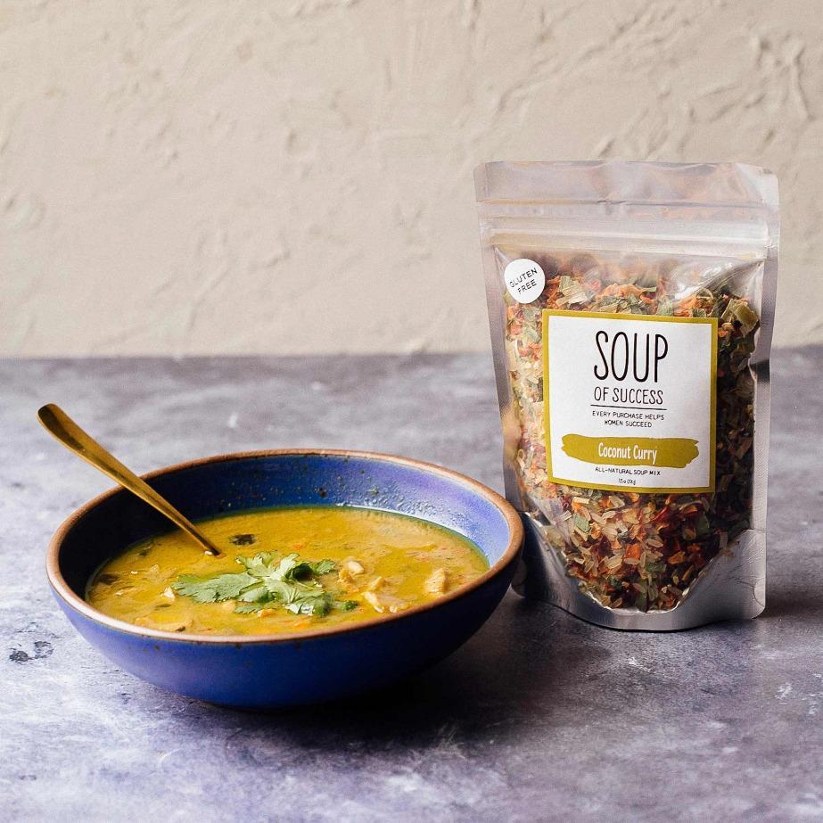 A blue bowl of soup on a marble surface beside a bag of Coconut Curry soup from the brand Soup of Success with a beige background