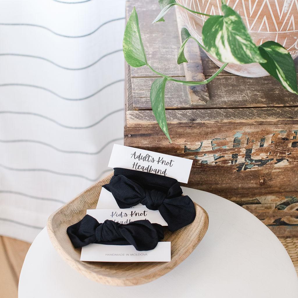 Two black knot headbands displayed on a wooden plate with a plant in the background.