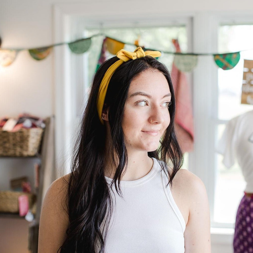 Young woman looking to the right with a mustard yellow knot headband in her dark hair in front of a colorful blurred background.