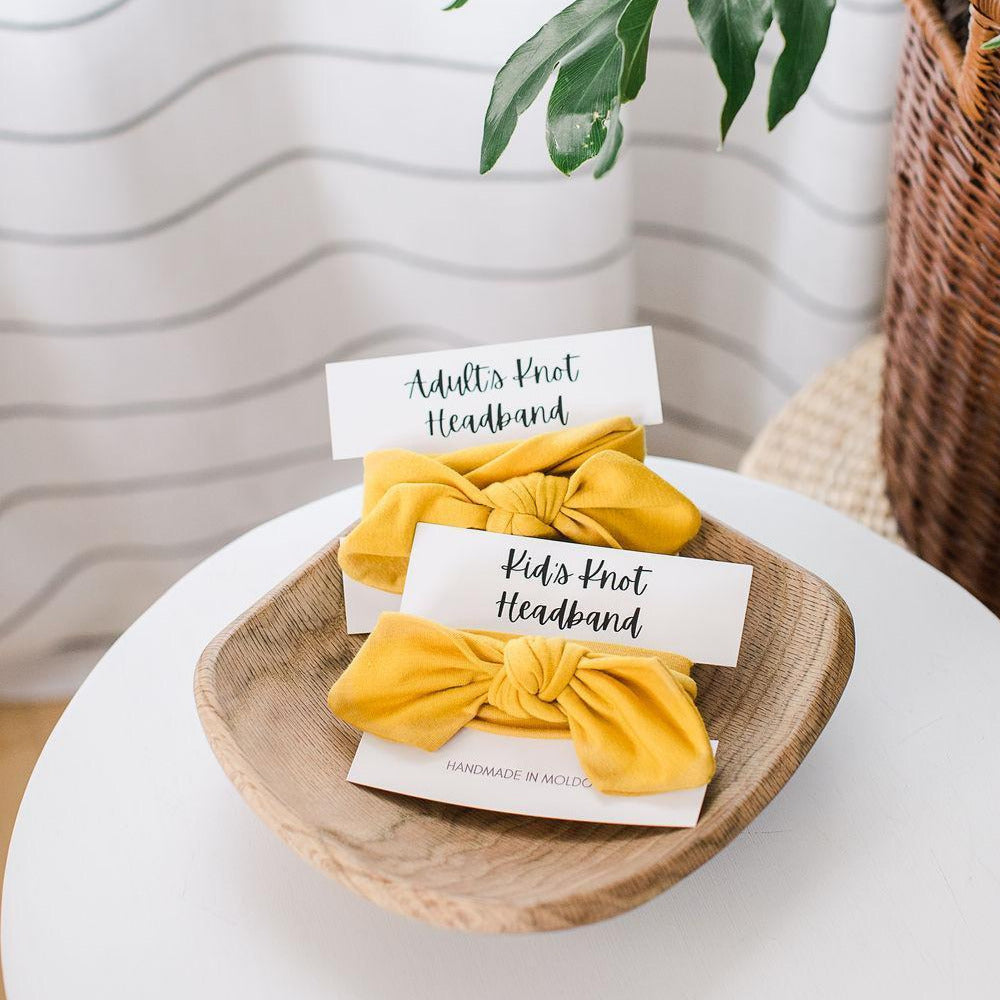 Two yellow knot headbands, one labeled for adults and the other for kids, placed on a wooden dish on top of a white surface with a striped background.