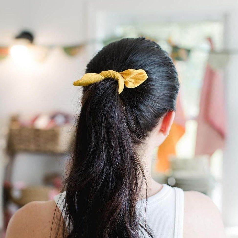 Long haired woman shot from behind with hair in a pony tail wearing a mustard yellow fabric hair tie.