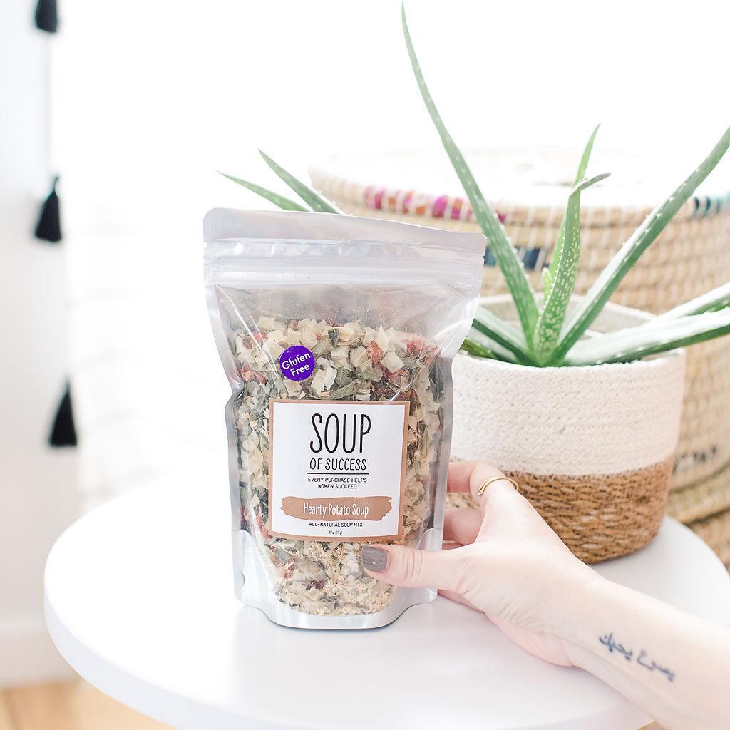 A packaged Hearty Potato Soup product from Soup of Success, held in front of a potted aloe vera plant.