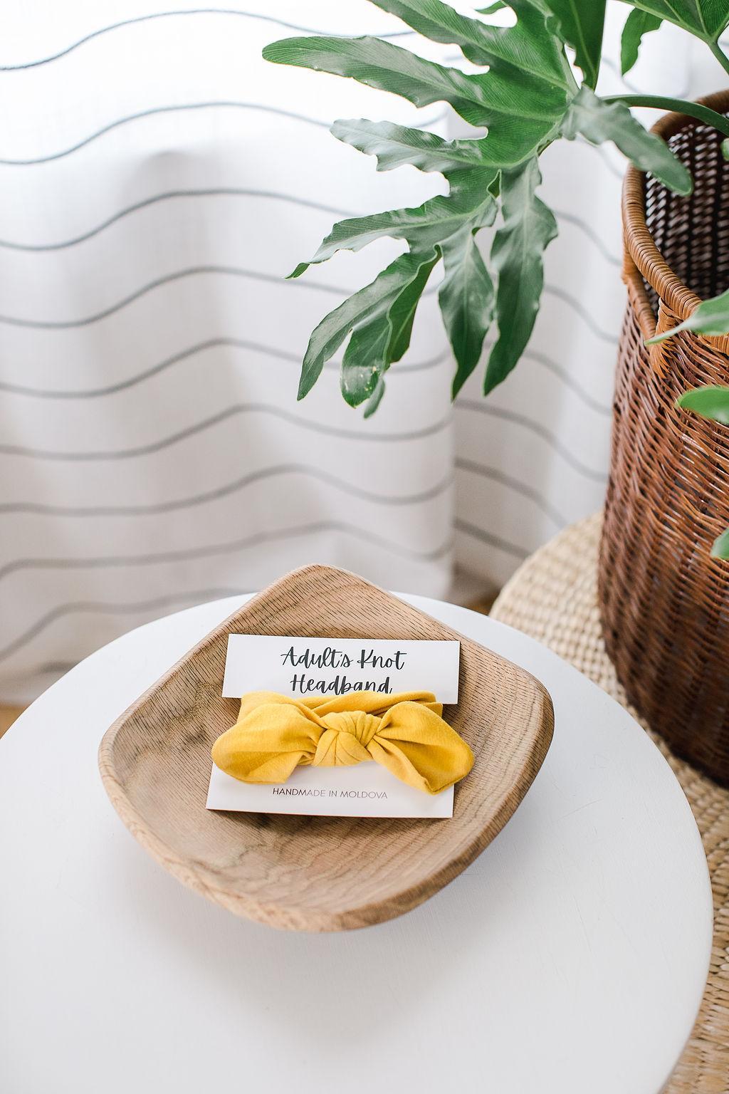 A yellow knot headband for adults on a white card sitting in a wooden bowl on a table with a plant to the side.