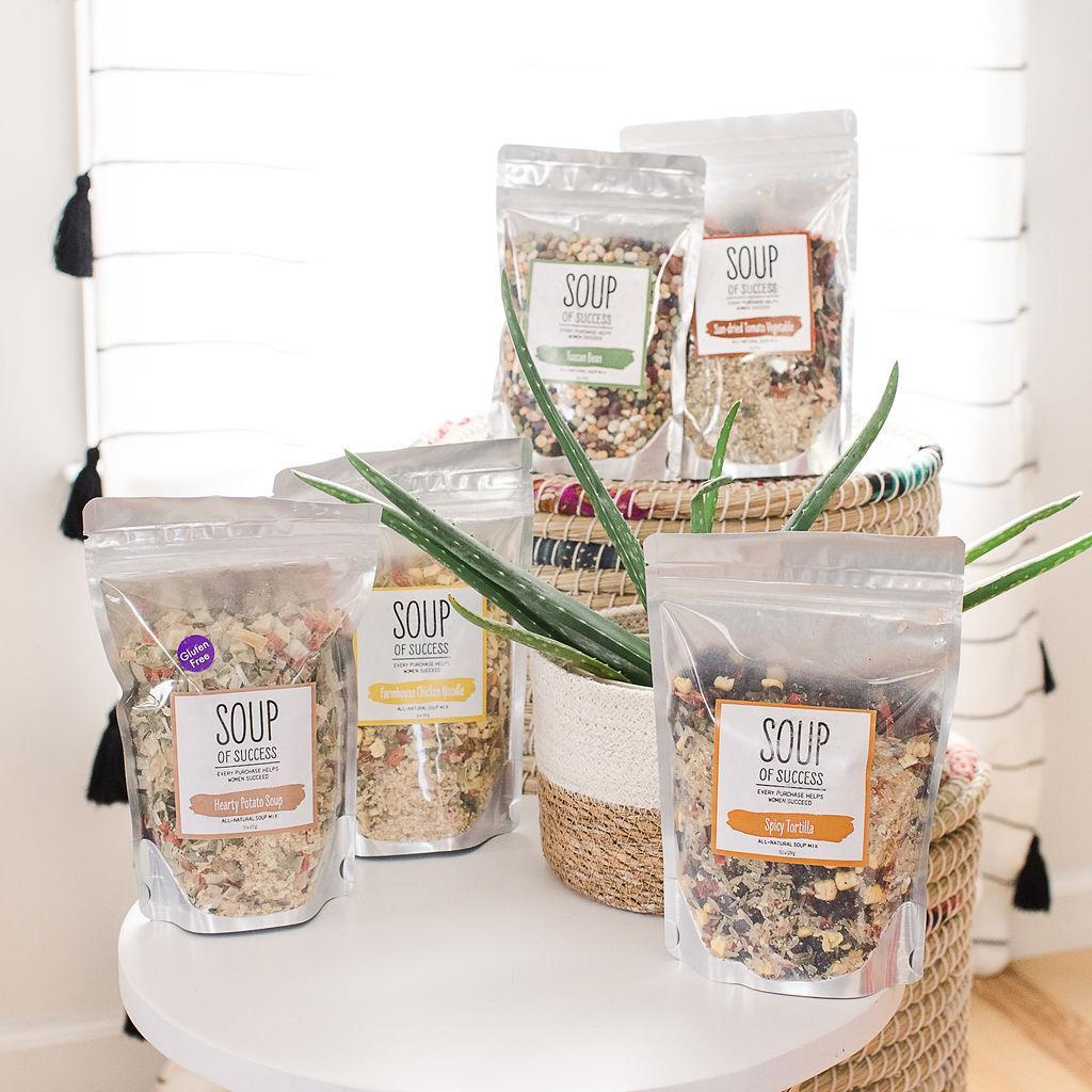 A collection of bags of soup mixes from the brand Soup of Success on a white table with a potted plant.