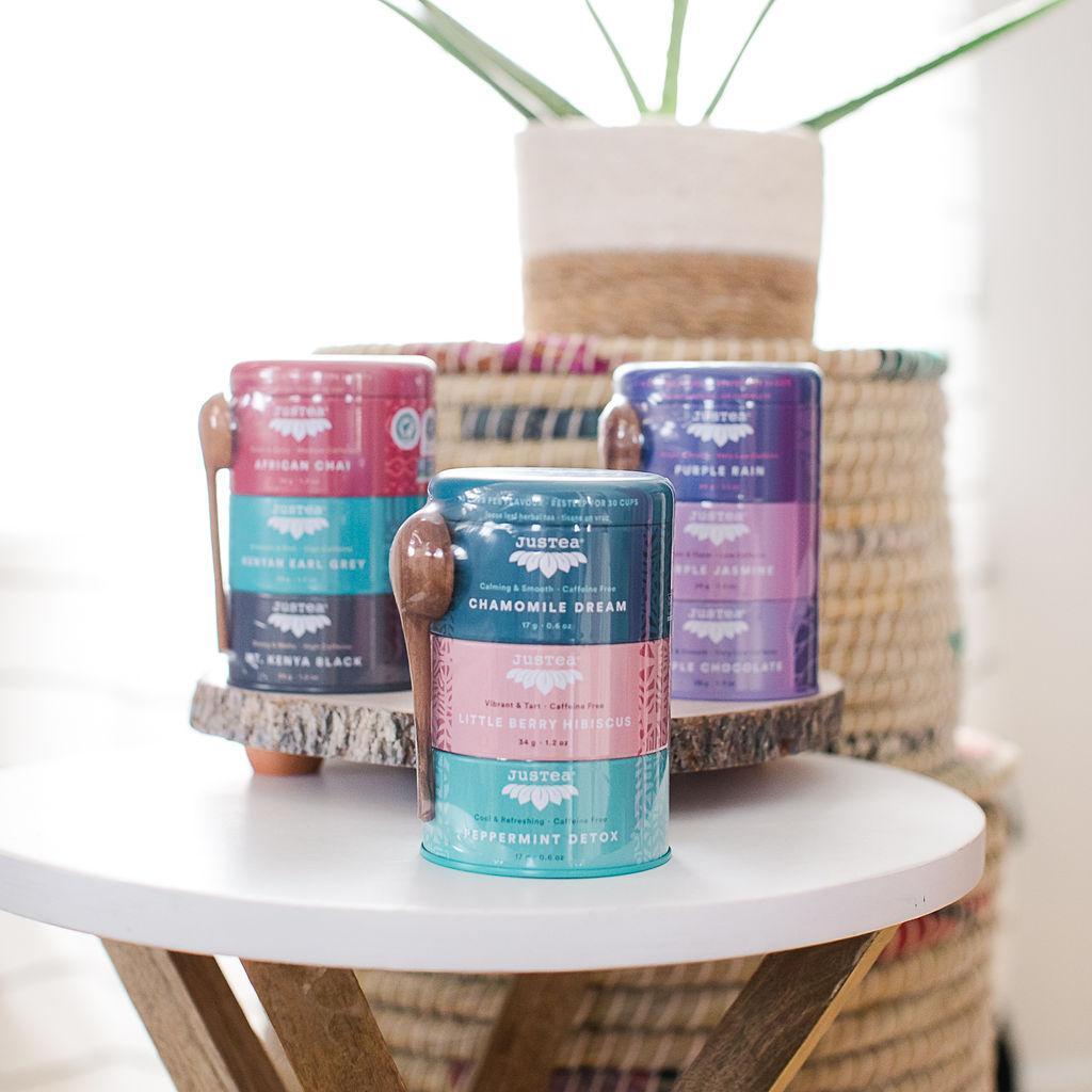 Three stacked tins of herbal tea with unique colors and labels for each variety, alongside a hand-carved Kenyan teaspoon, displayed on a table.
