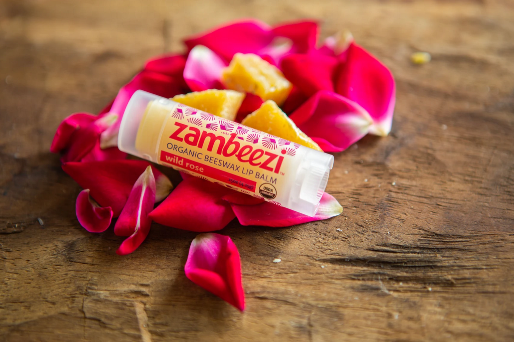Close up of a lip balm container on a wooden service with rose petals and wax beside it. Container says Zambeezi, organic beeswax lip balm, wild rose all in red.