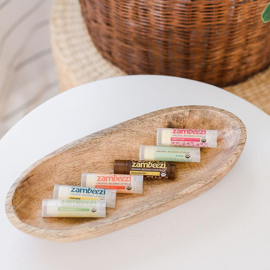 A collection of organic lip balms in various colors, displayed in an oval wooden tray, with the brand name &