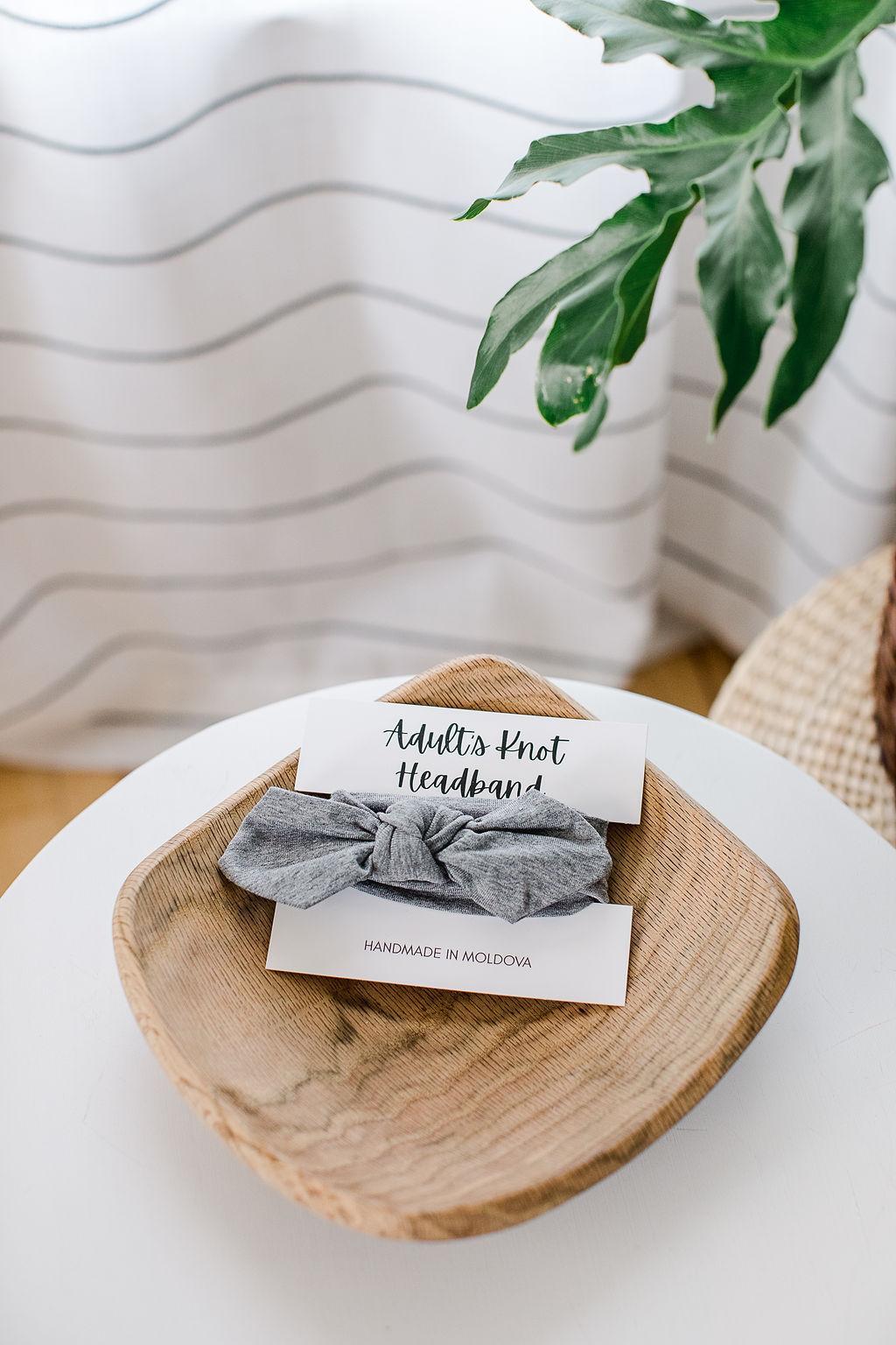 A heather gray knot headband for adults on a white card sitting in a wooden bowl on a table with a plant to the side.