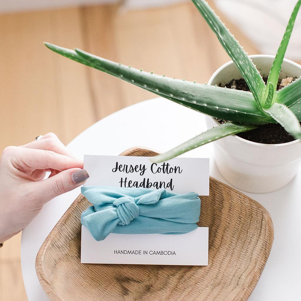 A light blue jersey cotton headband placed on a wooden platter, with a hand holding a cardboard tag indicating it is a handmade product from Cambodia.