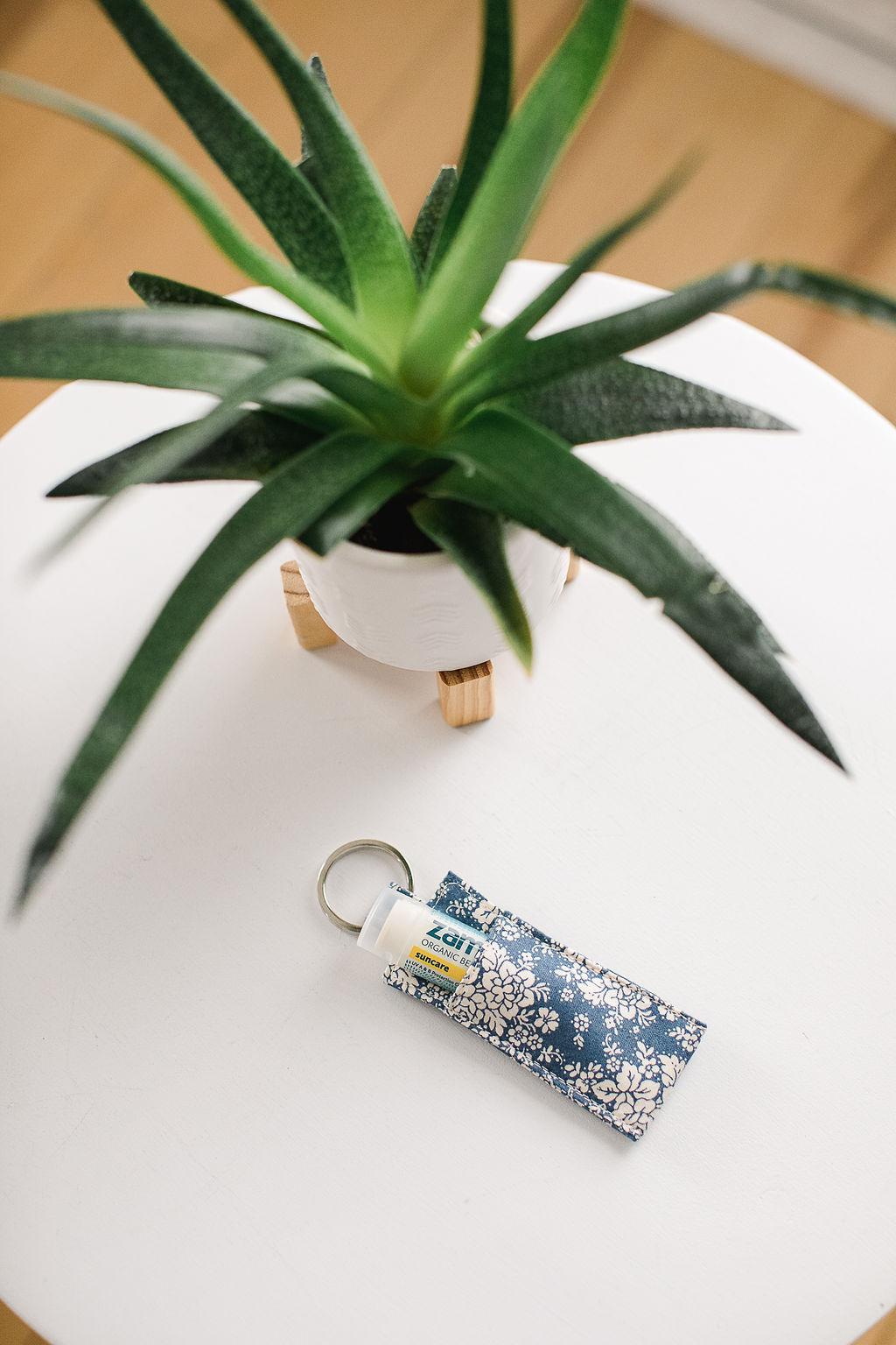 Lip balm with yellow and blue design in a blue floral fabric sleeve on a keychain sitting on a white table with a plant.