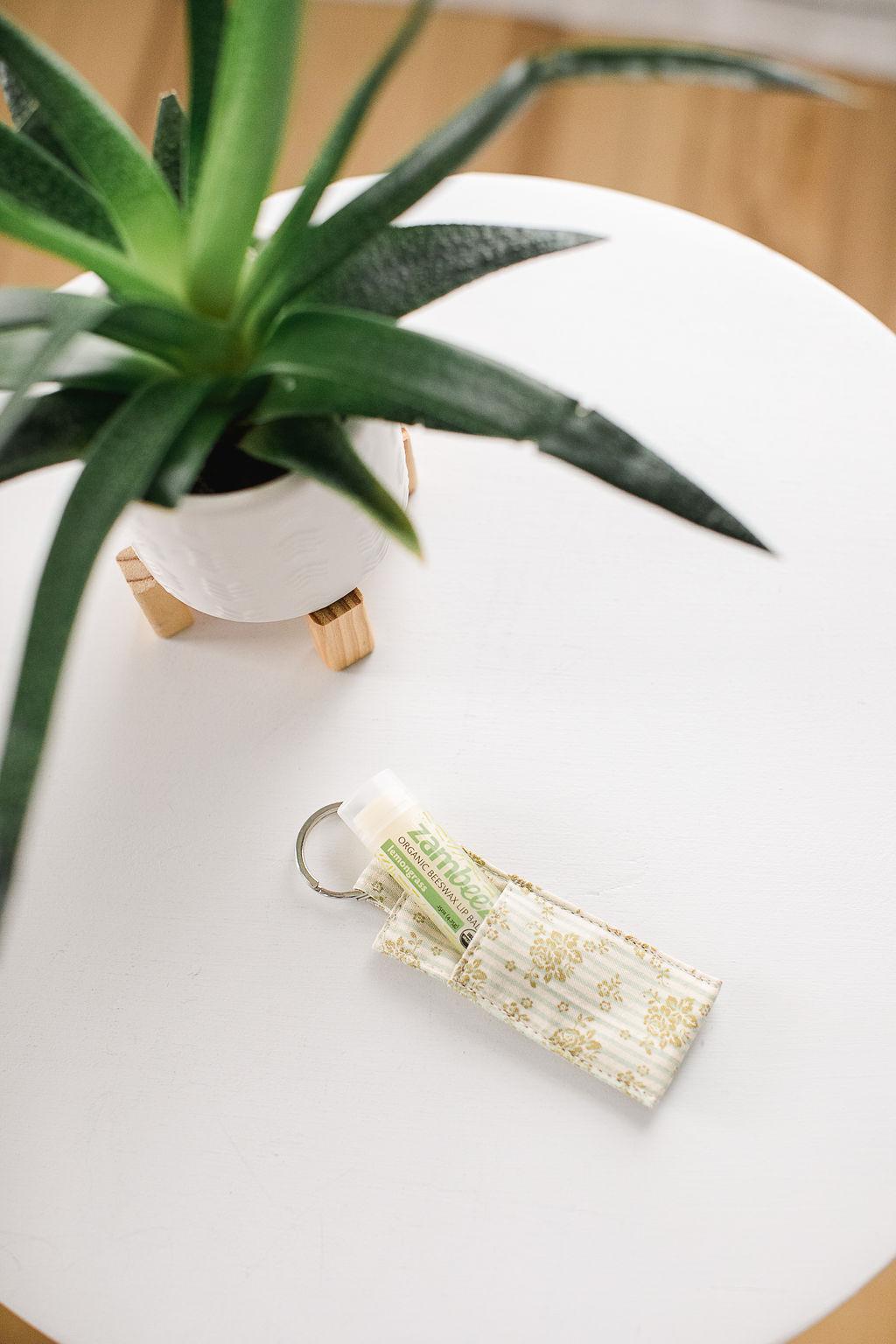 Lip balm with green design in a light colored fabric sleeve on a keychain sitting on a white table with a plant.