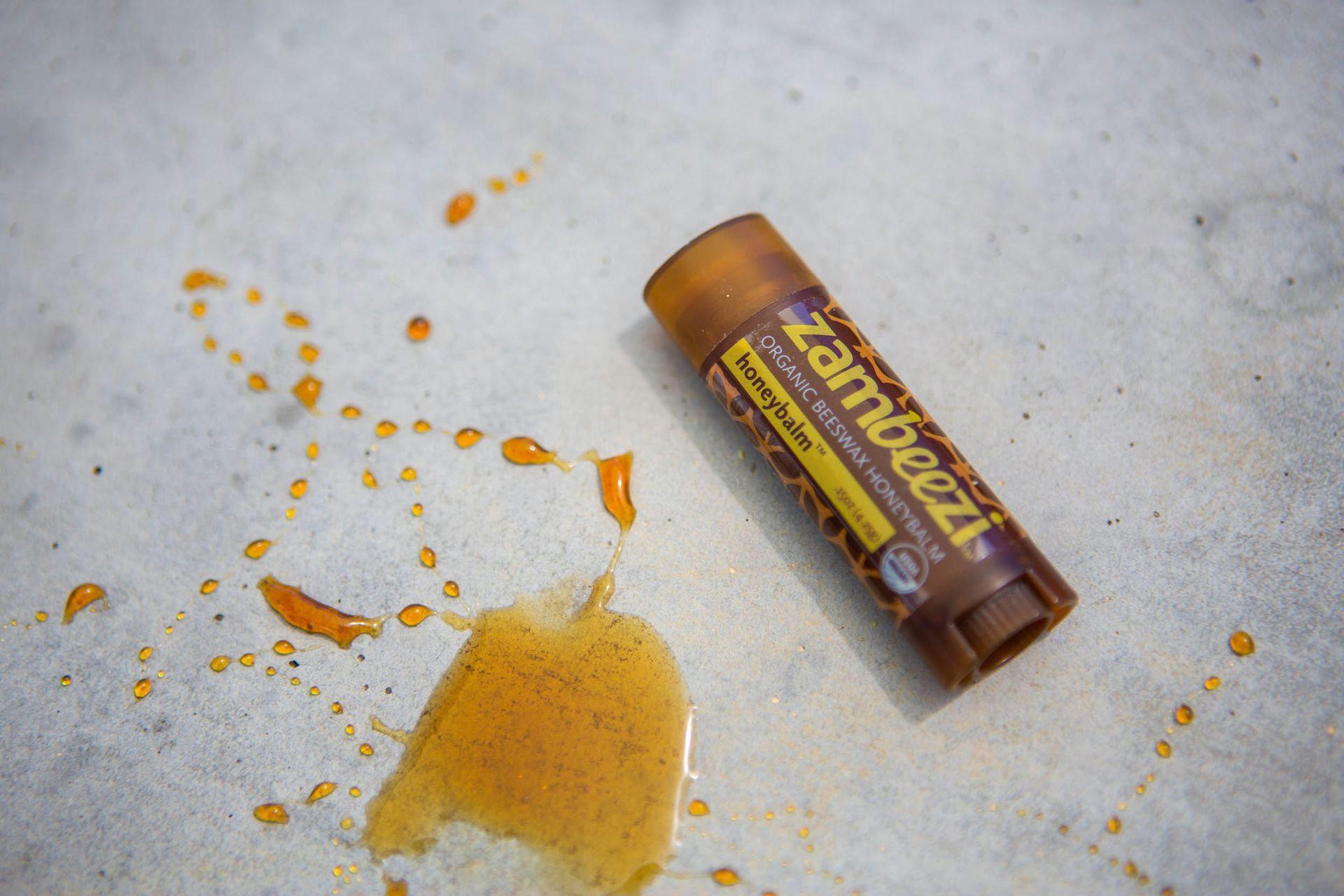 Close up of a brown lip balm container on a stone with honey drizzled beside it. Container says Zambeezi, organic beeswax honeybalm.
