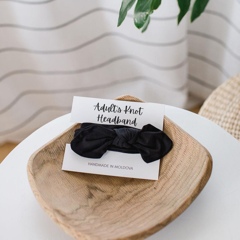 A black knot headband for adults on a white card sitting in a wooden bowl on a table with a plant to the side.