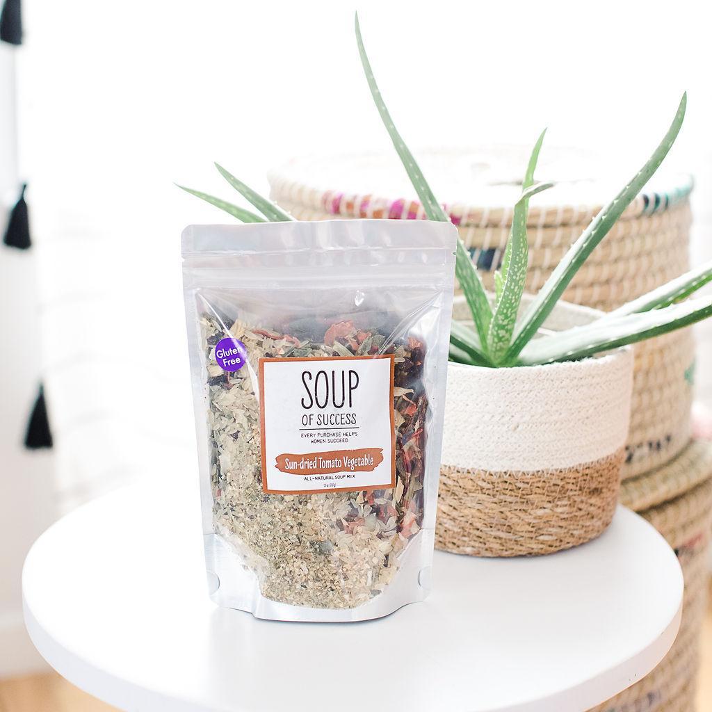 A package of Sun-dried Tomato Vegetable Soup from Soup of Success sitting on a white table with a potted plant behind.