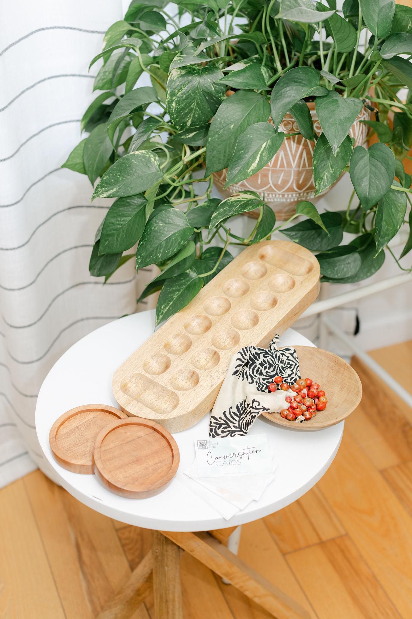 Round white table with a wooden mancala board, wooden coasters, and a plant in the background.