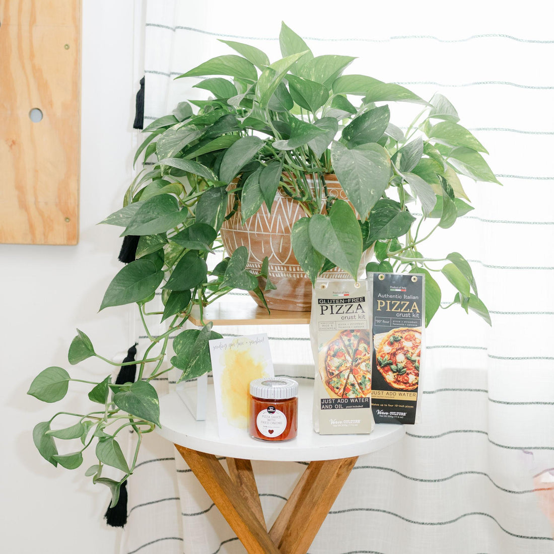 A potted green pothos plant sits on a small white side table next to packaged pizza toppings and a small jar, with a striped curtain and light wood wall in the background.