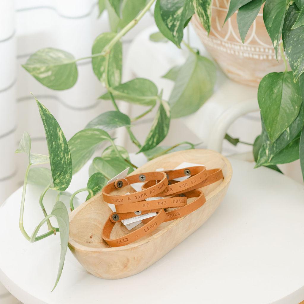 Wooden dish with a collection of light brown leather bracelets featuring different sayings on a white table with a plant in the background