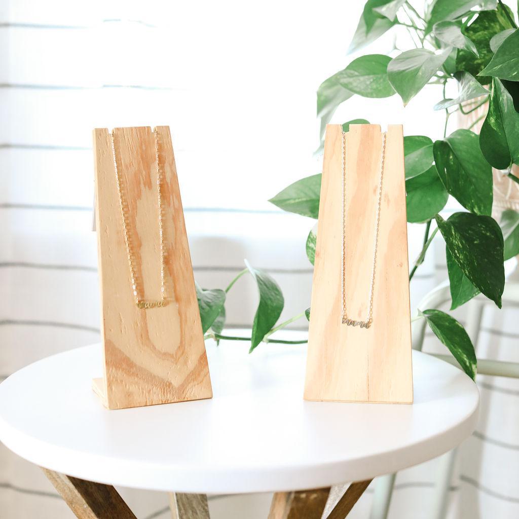 Two wooden jewelry stands with necklaces on a white surface with greenery in the background