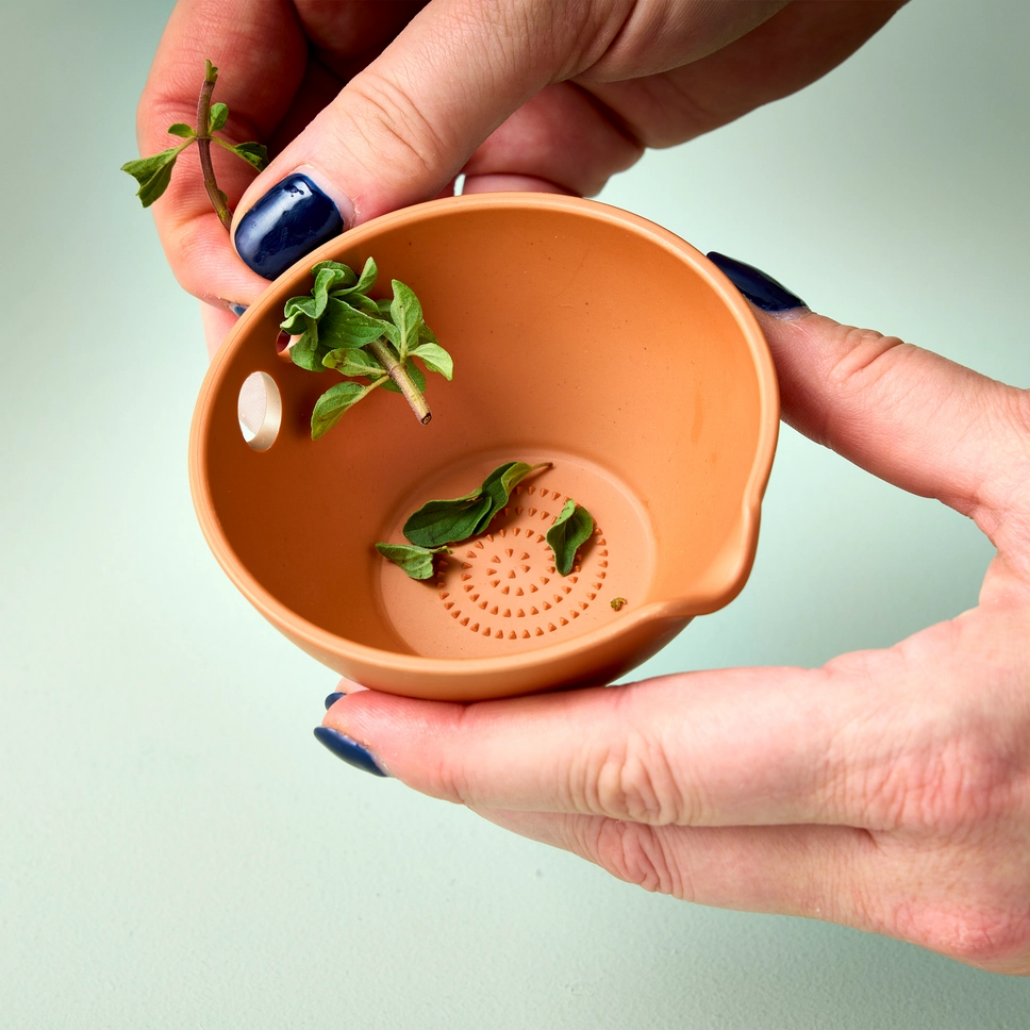 Person holding a small terracotta colored bowl with holes, stripping leaves off herbs on a light green background
