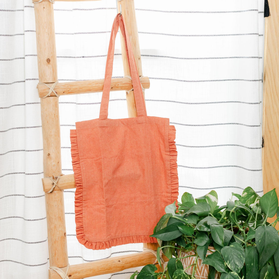 Rust colored ruffle-edge tote bag hanging on a wooden ladder with a white striped wall in the background