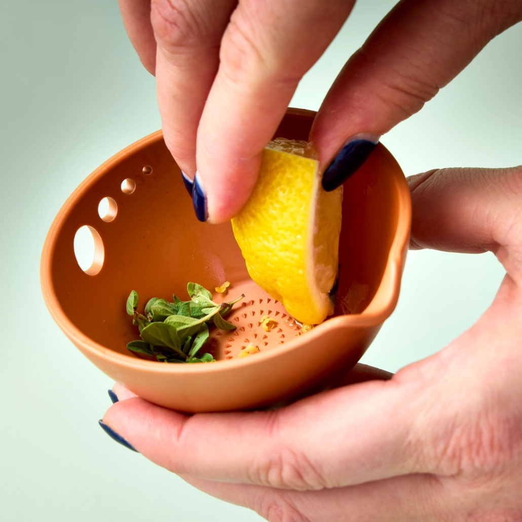 Person holding a terracotta colored bowl with a lemon and herbs against a light green background