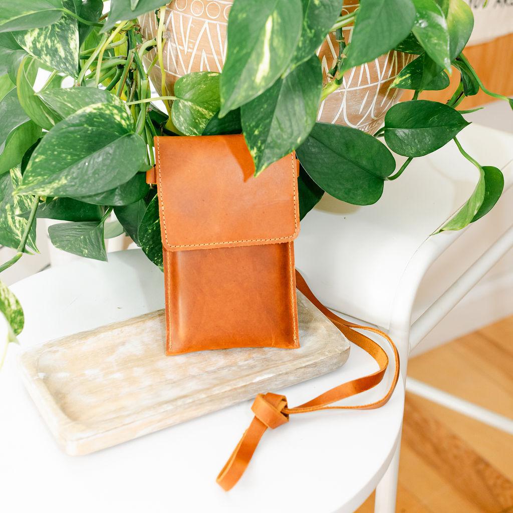 A brown satchel bag with a shoulder strap placed on a white surface, partially hidden by green leafy plants in the background.