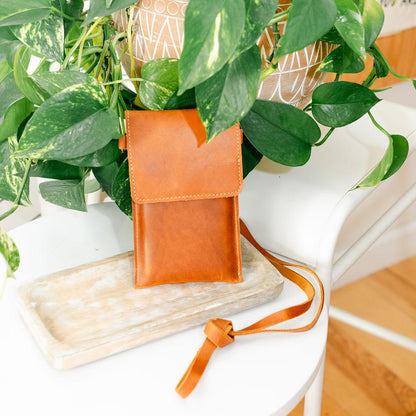 A brown satchel bag with a shoulder strap placed on a white surface, partially hidden by green leafy plants in the background.