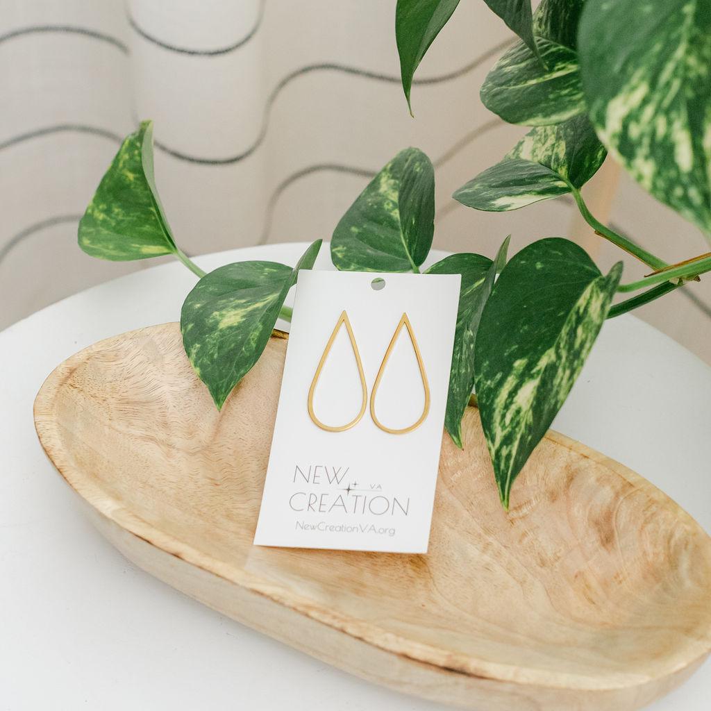 A pair of large teardrop post earrings made of brass, displayed on a wooden dish with a plant in the background.