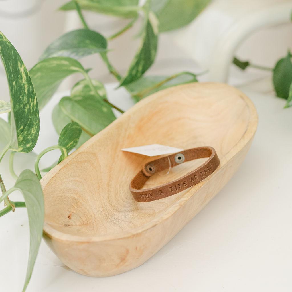 Wooden dish with leather bracelet featuring the saying "such a time as this", surrounded by green leaves on a light background
