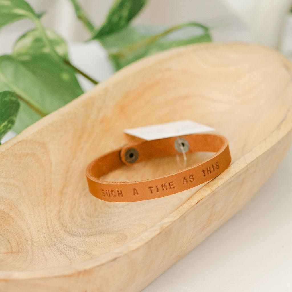 Close up of a wooden dish with a lighter leather bracelet featuring the saying "for such a time as this", surrounded by green leaves on a light background