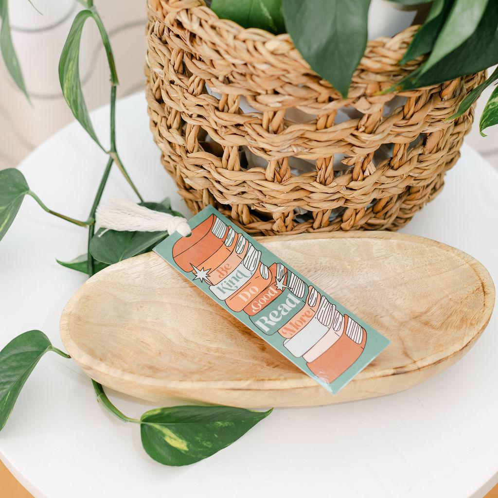 A colorful bookmark with text, placed on a wooden surface with a woven basket and green leaves in the background.