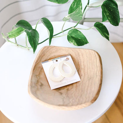 Wooden dish with a card featuring white woven hoop earrings on a white surface with green leaves.