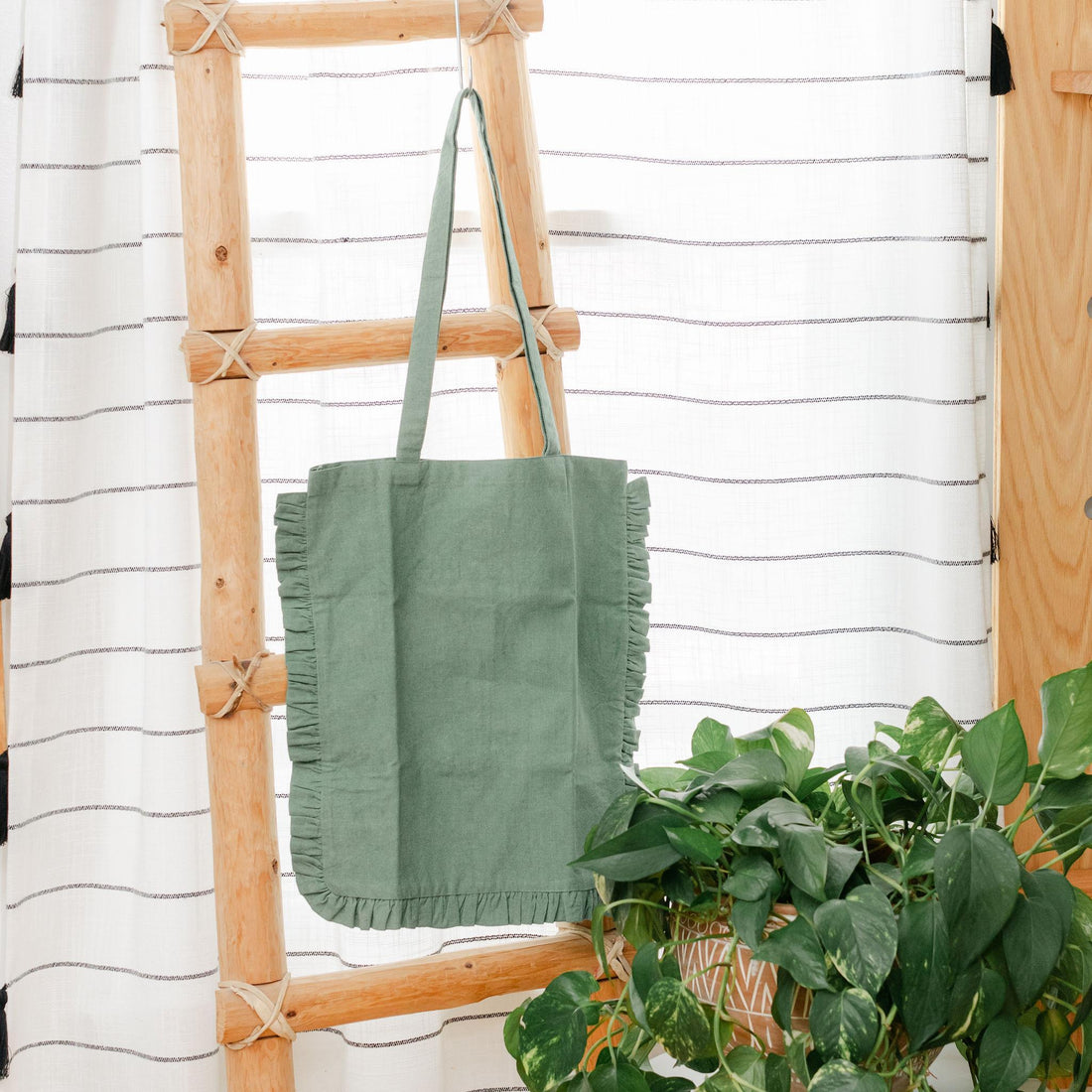 Green ruffle-edge tote bag hanging on a wooden ladder with a plant in the foreground against a striped wall.