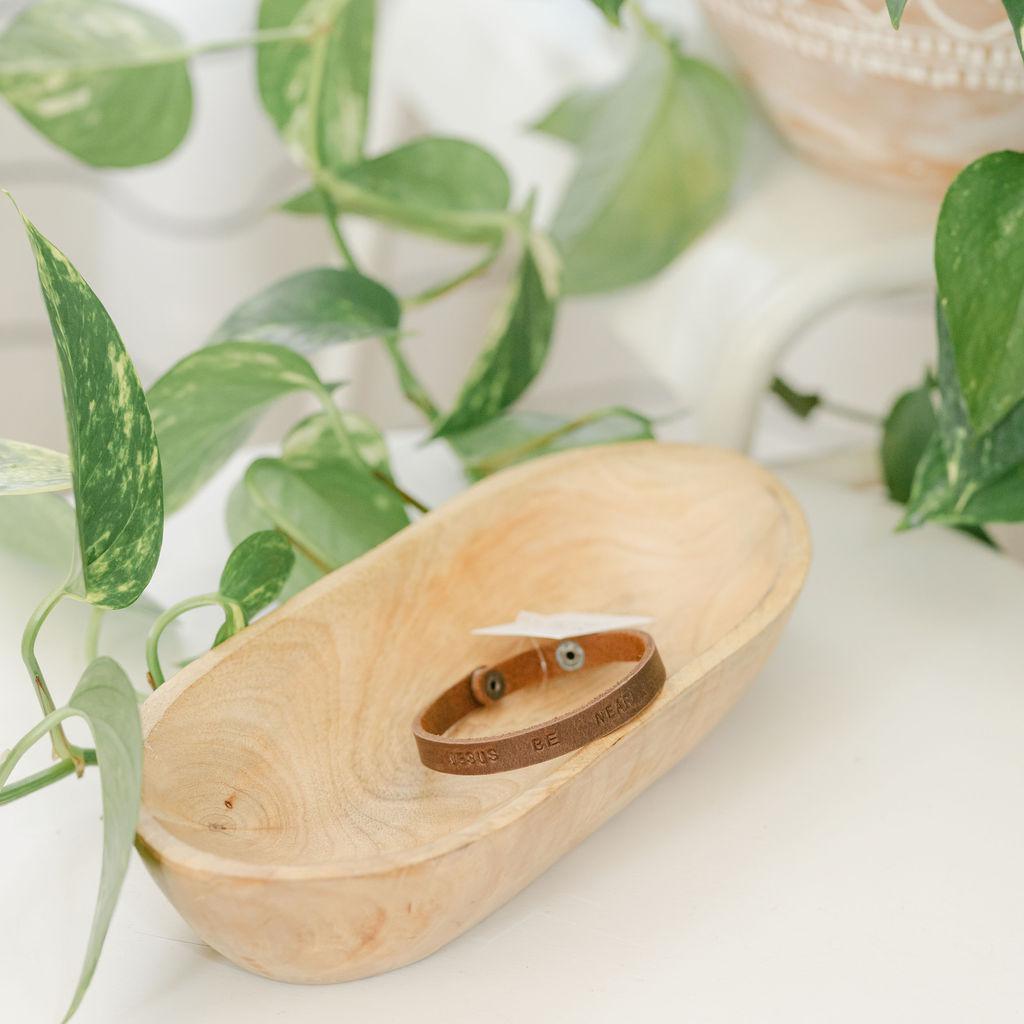 Brown leather bracelet in a wooden dish with green leaves in the background