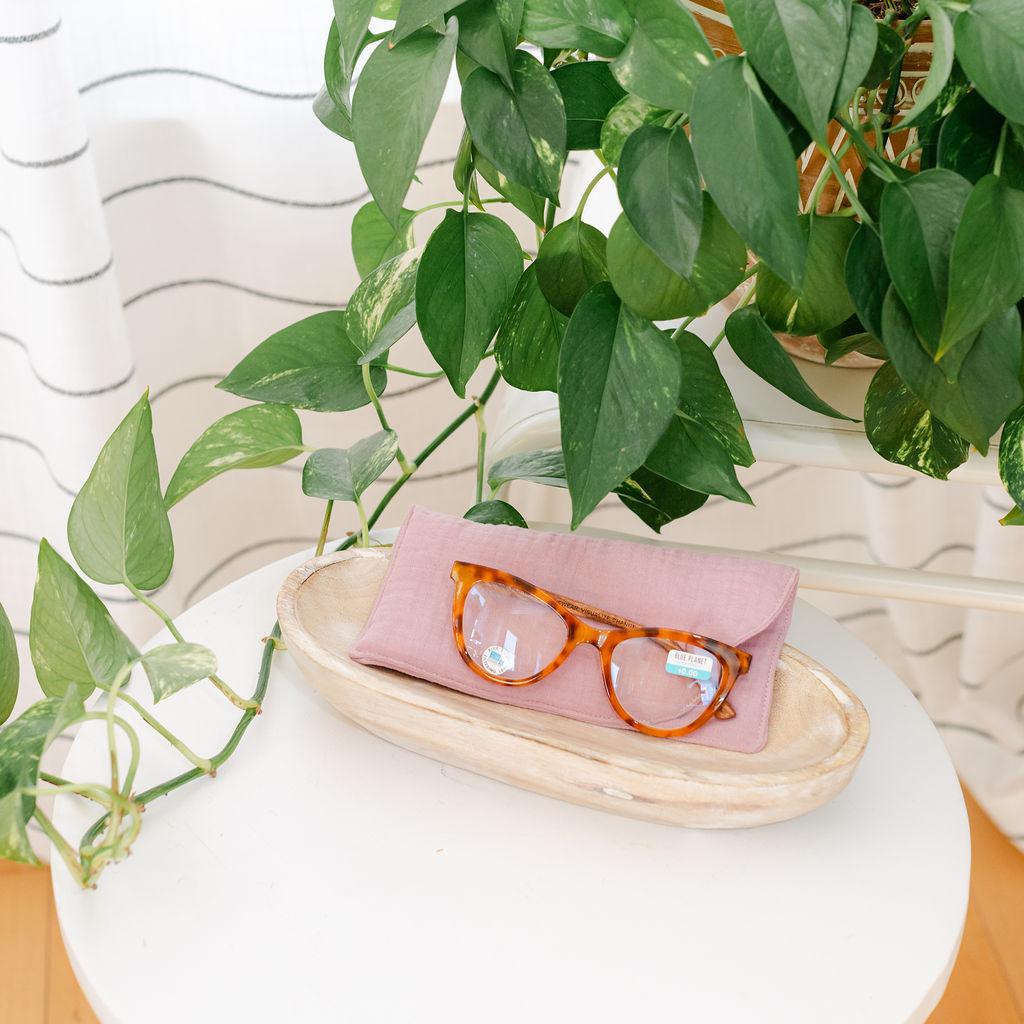A pink muslin sunglass case w/ sunglasses displayed on a wooden tray with a plant in the background.