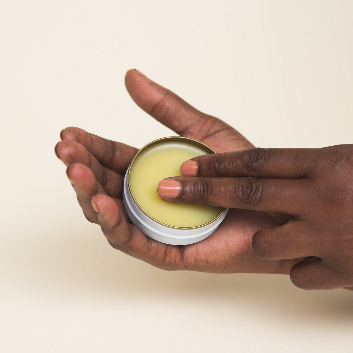 Hand applying balm from a small jar on a beige background