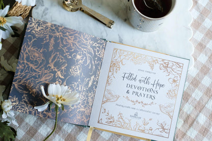 Open book with decorative cover and text on a checkered tablecloth with flowers and a cup.