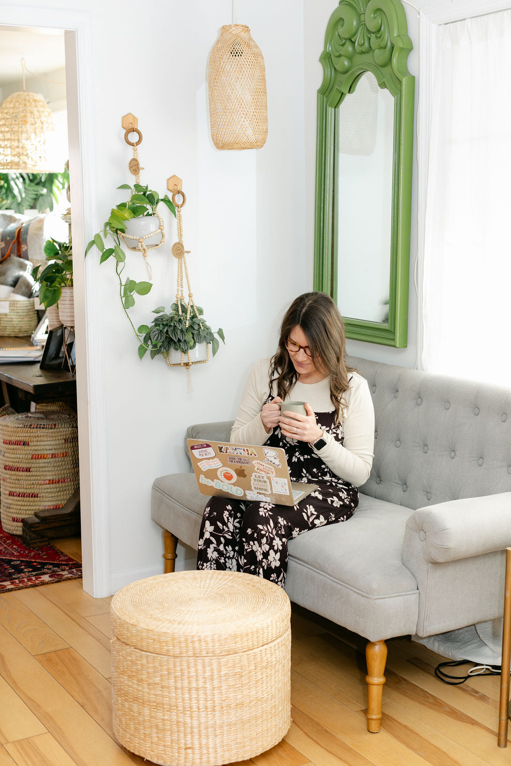 Woman sitting on a gray sofa in a cozy corner with plants and a green mirror.