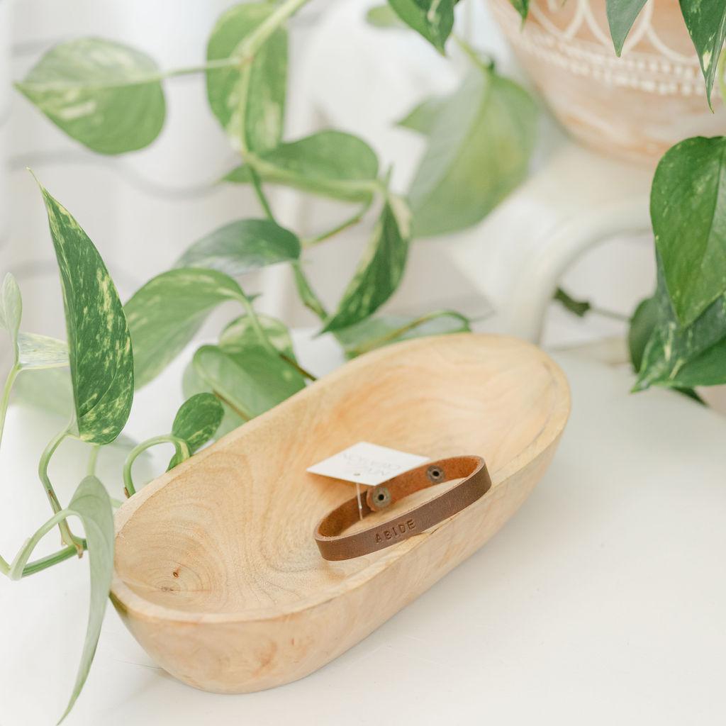 Wooden dish with a brown leather bracelet on a white surface with green leaves in the background