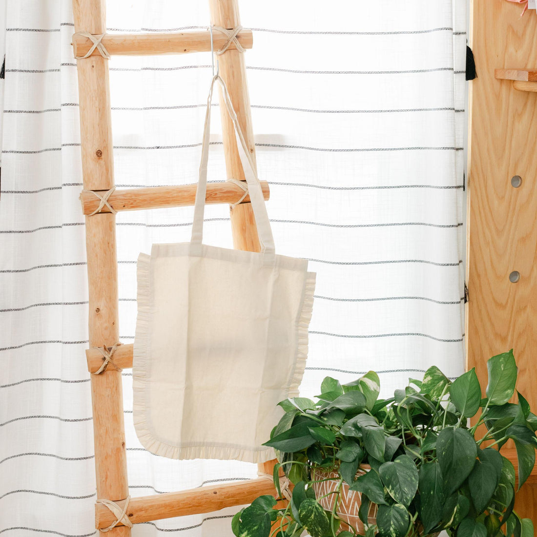 Cream ruffle-edge tote bag hanging on a wooden ladder with a plant in the foreground against a striped wall.