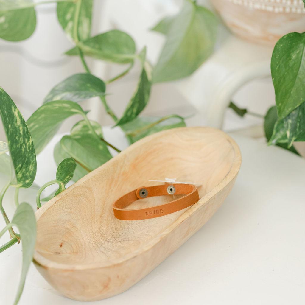 Wooden dish with a light brown leather bracelet on a white surface with green leaves in the background