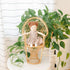 A handmade plush doll with brown buns wearing a floral dress with a plant in the background