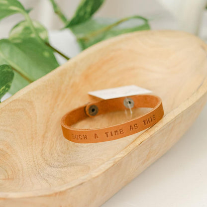 Light brown leather bracelet with engraved text on a wooden surface with a plant in the background