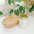 A candle with white labels, set on a white surface with green leaves in the background.