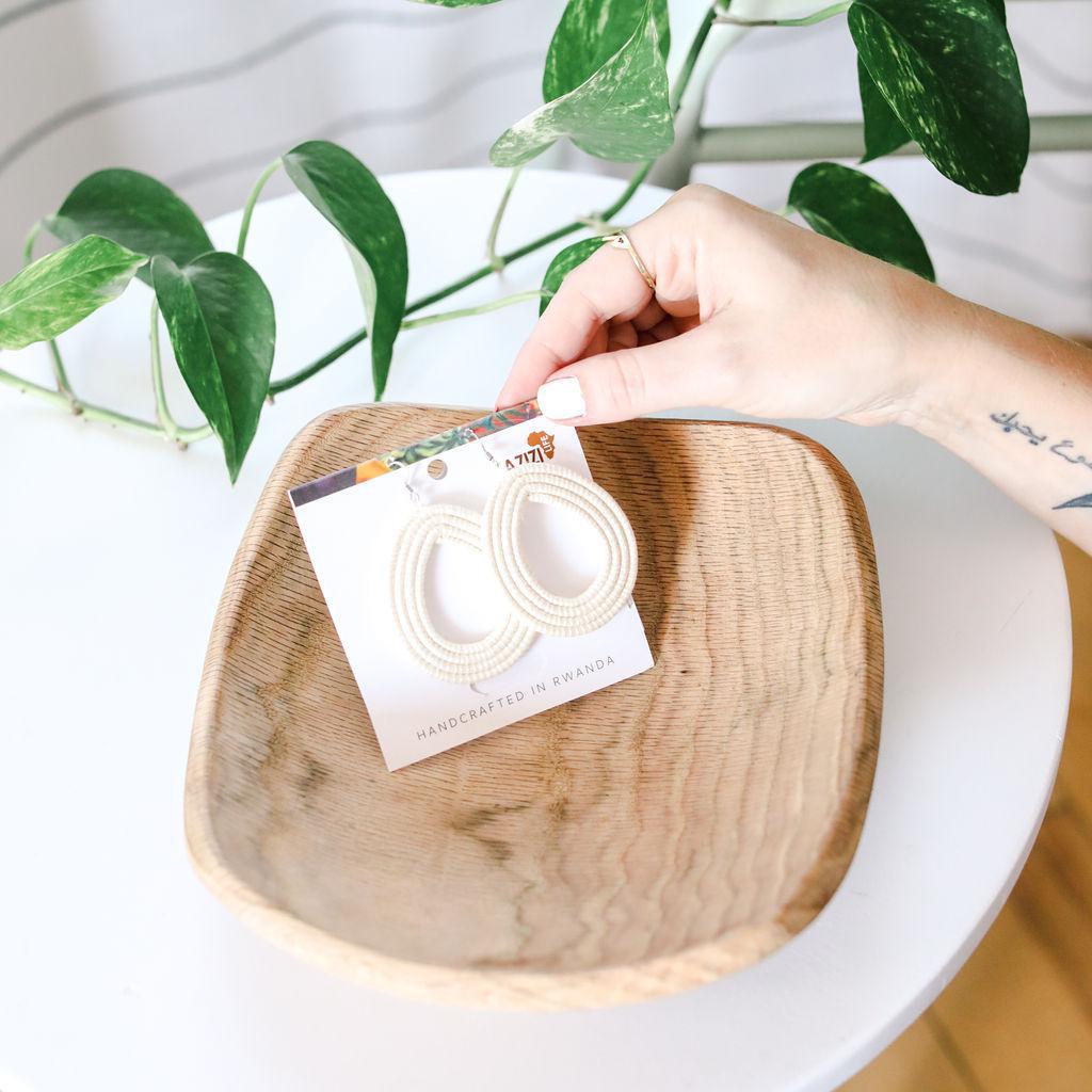 Hand holding a product card featuring white woven hoop earrings over a wooden bowl with green leaves in the background
