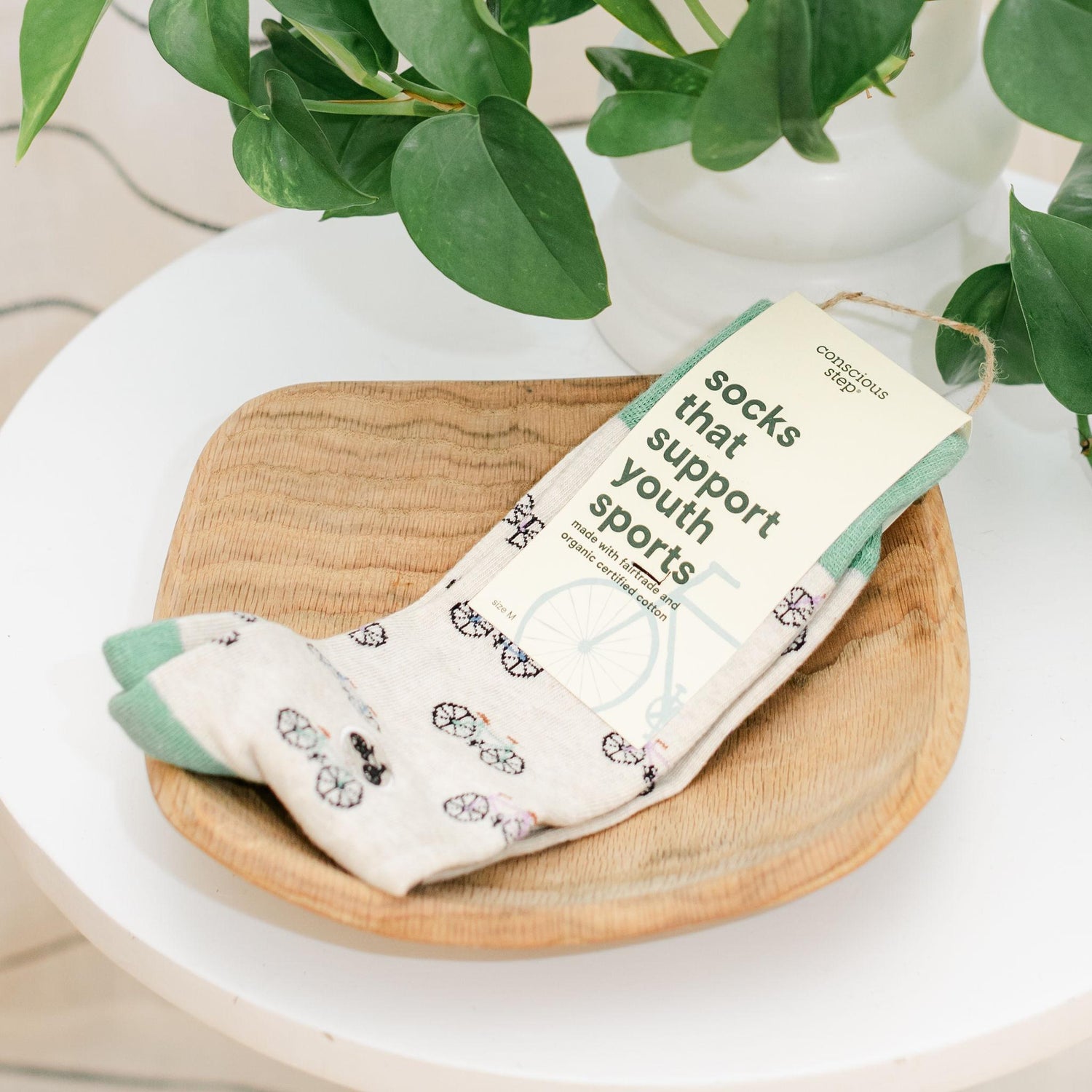 Pair of light green socks featuring bicycles with a packaging label on a wooden tray with a plant in the background